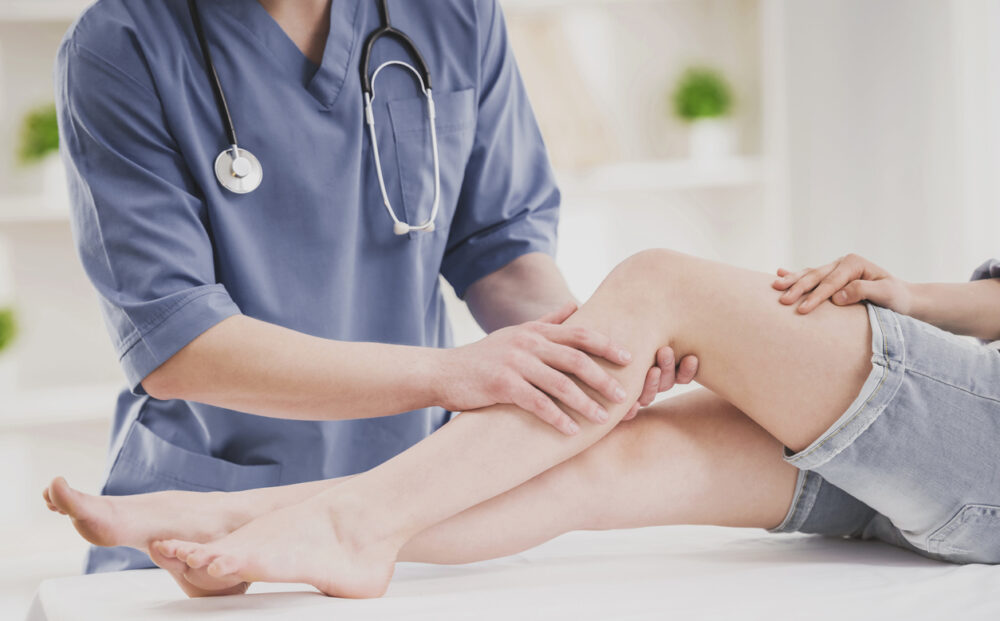 Close up. Doctor in Blue Uniform Comfforting Leg of Sitting Young Woman in Doctor Office. Traumatology Medical Devices. Medical Treatment in Clinic and Healthcare Professional Concept.