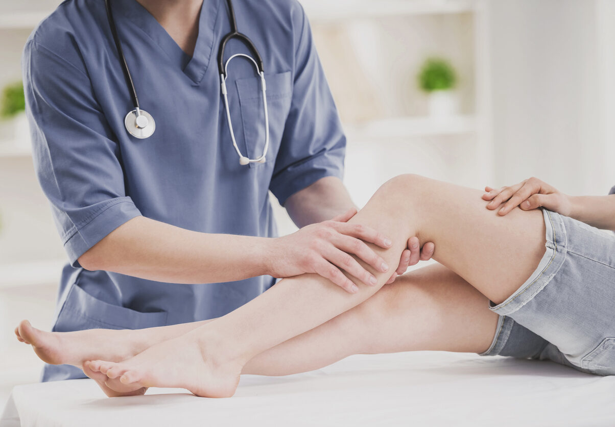 Close up. Doctor in Blue Uniform Comfforting Leg of Sitting Young Woman in Doctor Office. Traumatology Medical Devices. Medical Treatment in Clinic and Healthcare Professional Concept.